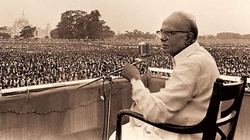 Sepia photograph of Jaya Prakash Narayan seated at a microphone on an open-air dais, addressing an enormous crowd filling a maidan, with a large domed building in the distance
